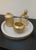Three gold decorative home decor items, including a ribbed vase, a small bowl, and a seated figurine, arranged on a round white tray with a gold rim, set on a black surface against a beige wall