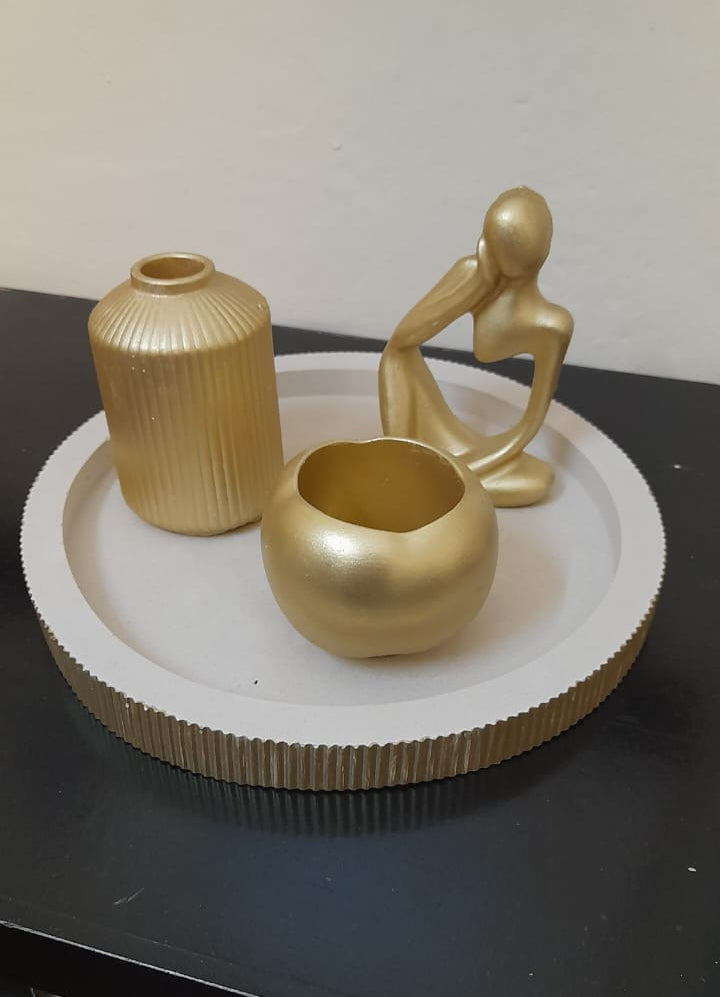 Three gold decorative home decor items, including a ribbed vase, a small bowl, and a seated figurine, arranged on a round white tray with a gold rim, set on a black surface against a beige wall