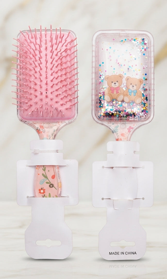 Two white hairbrushes with pink floral handles, one featuring pink bristles and a pink bear pattern, and the other with a clear liquid and multicolor glitter container with cute bear illustrations, set against a marble background.