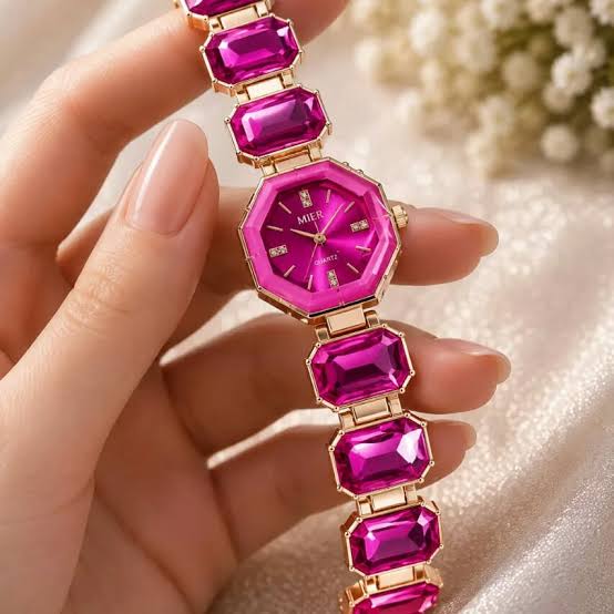 A close-up of a hand holding a gold-tone watch with bright magenta pink gemstone links and a matching pink dial. The watch has a hexagonal face with gold-tone markers and hands.