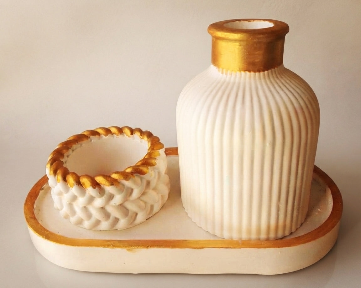 A cream-colored ceramic set featuring a ribbed bottle with a gold-painted neck and a matching textured bowl with a gold braided rim, both resting on a coordinating oval tray.