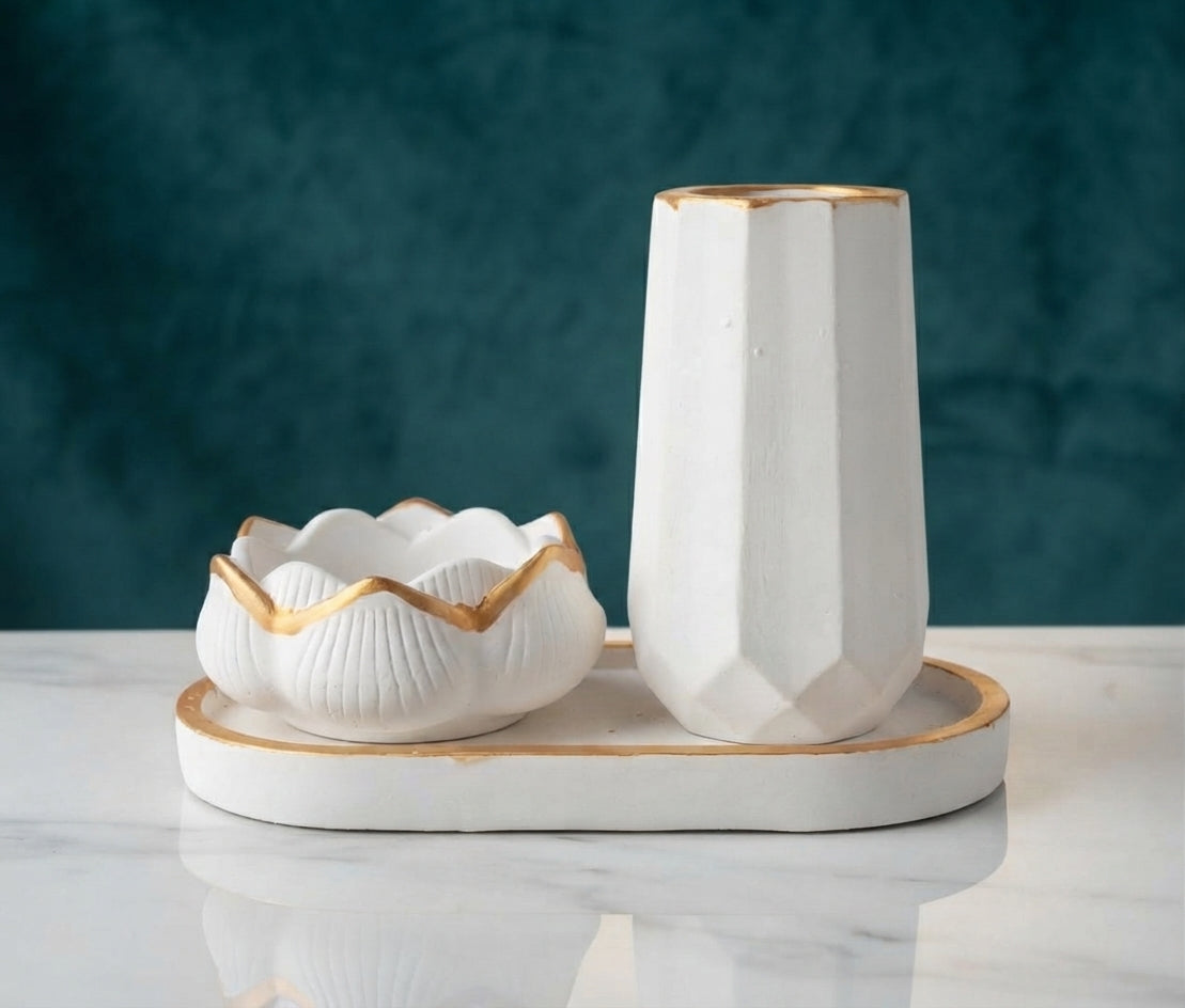 White ceramic tableware set with gold-painted edges, including a tall faceted cylinder vase, a shallow scalloped bowl, and an oval tray, displayed against a textured gray concrete background.