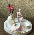 A decorative white and pink table set with gold leaf accents, featuring a ribbed vase, pink thinker figurine, jewelry jar, and round tray by Zeeno Place.