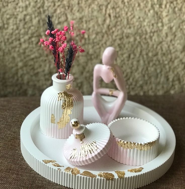 A decorative white and pink table set with gold leaf accents, featuring a ribbed vase, pink thinker figurine, jewelry jar, and round tray by Zeeno Place.
